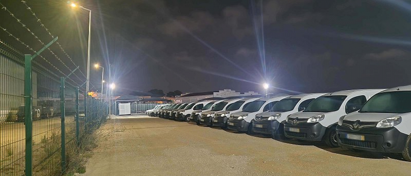 Car park lit at night, with vehicles parked in a row along the fence