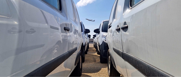 Viaturas estacionadas no parque, com um avi&atilde;o a levantar ao fundo